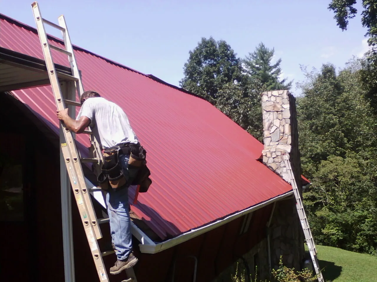 Roofer on a ladder beside a red metal roof and stone chimney — Soffit & Fascia Repair by Roofing Brothers in Forks